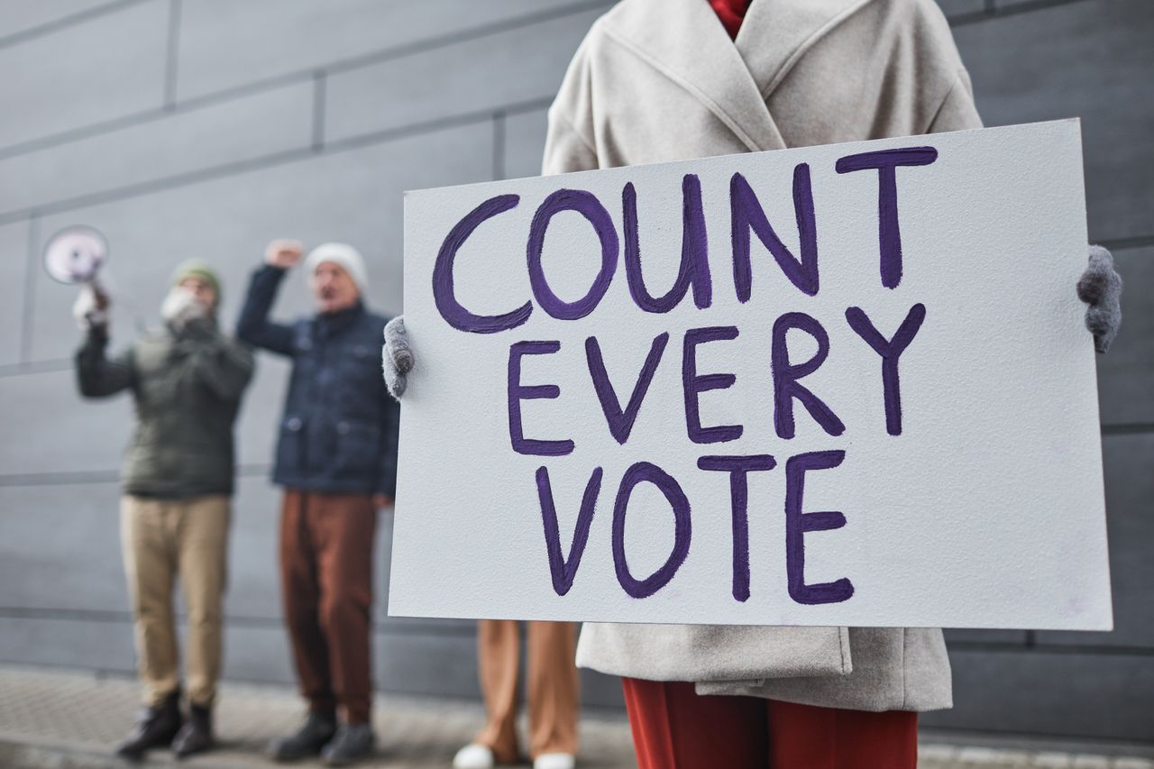 creative-01 Protesters holding signs and chanting for fair vote count during a winter rally.