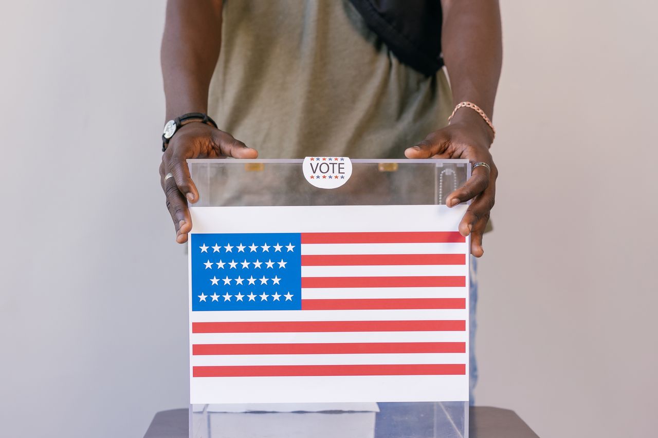 digital-01 Close-up of a person placing a vote in a transparent ballot box with an American flag print.