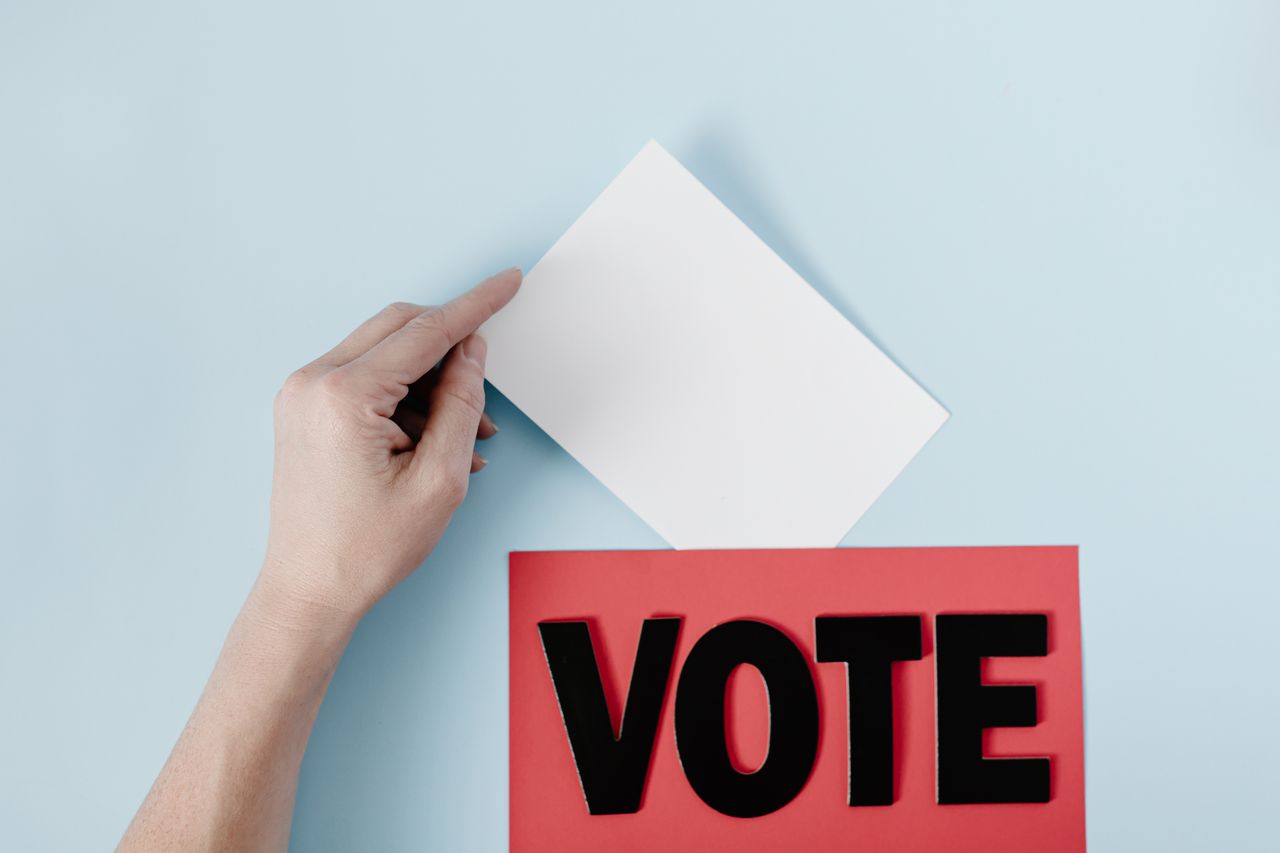 creative-03 A hand places a ballot into a red box with the word VOTE against a blue background.
